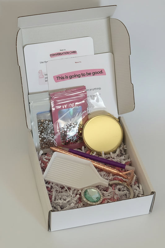An open white gift box filled with assorted stationery-style gift items, including a gold compact, pink and champagne rhinestones in bags, a purple rhinestone picker, a green gem purse hanger, and printed cards inside shredded paper filler.