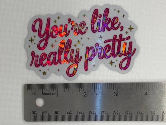 A die-cut vinyl sticker with the phrase 'You're like, really pretty' in holographic pink and purple glitter script, shown next to a metal ruler for scale on a white textured background.