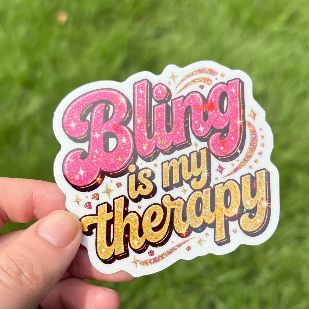 A hand holding a die-cut sticker that reads 'Bling is my therapy' in pink and yellow bubble letters with a white border, set against a blurred green grass background.