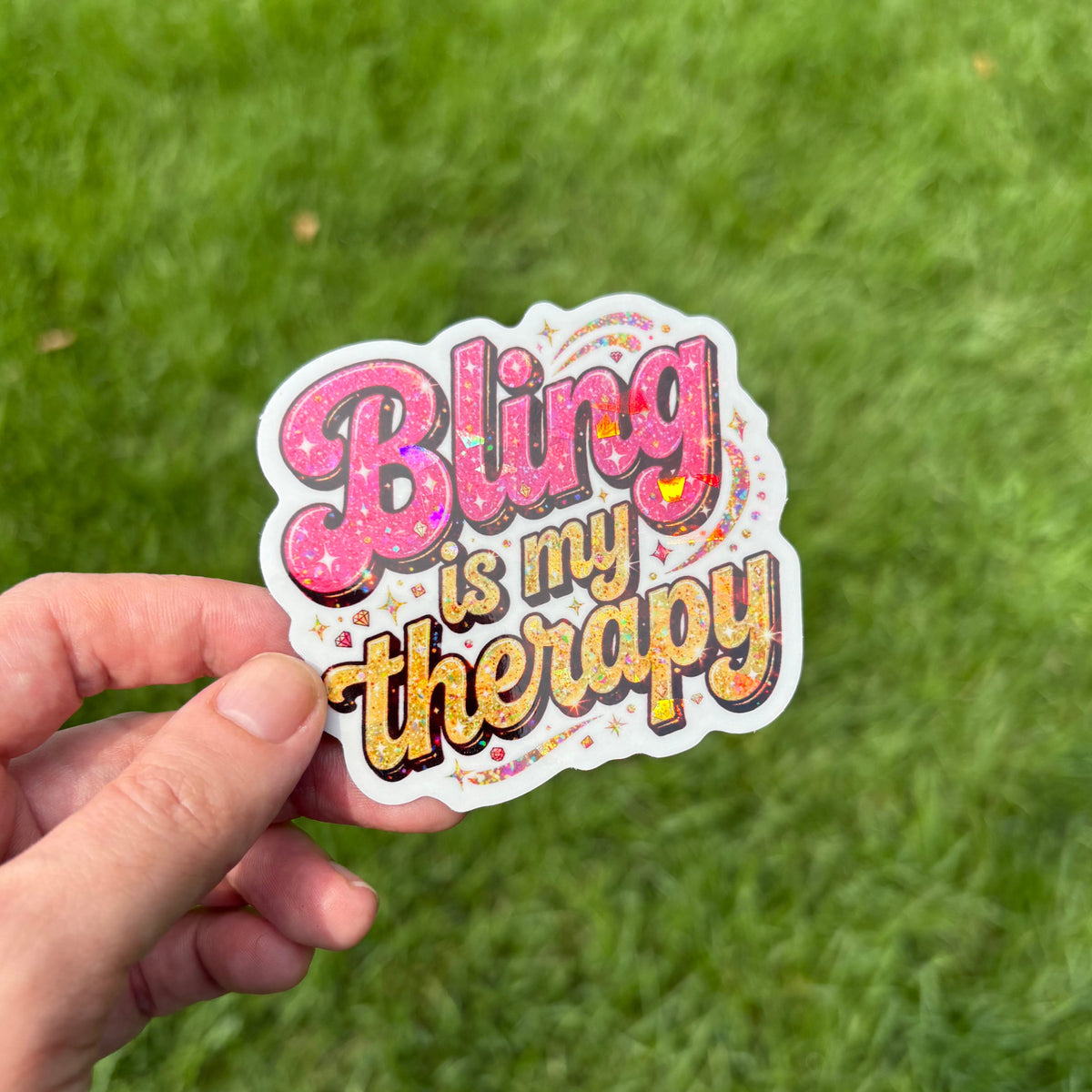 A hand holding a die-cut sticker that reads 'Bling is my therapy' in pink and yellow bubble letters with a white border, set against a blurred green grass background.
