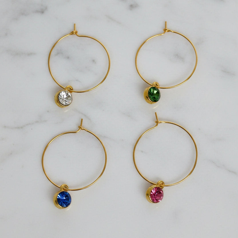 Four gold hoop wine markers arranged in a 2x2 grid on a textured white background. Each hoop has a tiny round gemstone dangling below: clear, green, blue, and pink.