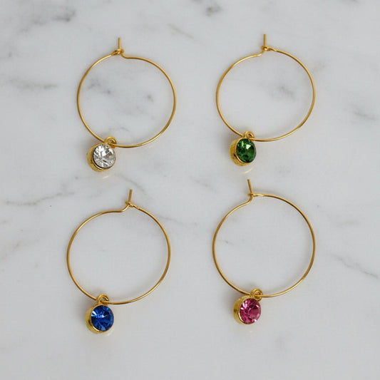 Four gold hoop wine markers arranged in a 2x2 grid on a textured white background. Each hoop has a tiny round gemstone dangling below: clear, green, blue, and pink.