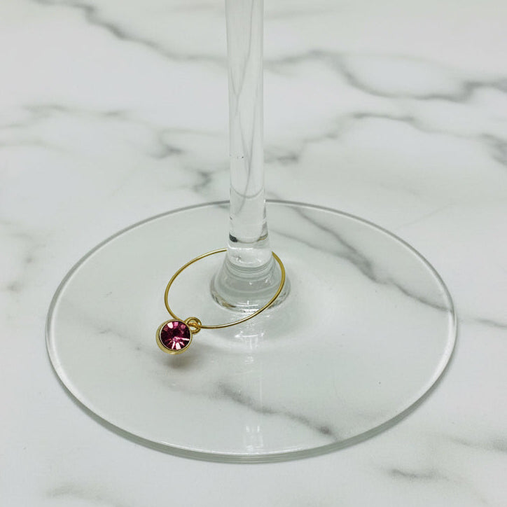 A gold wire wine glass marker with a small round pink gemstone bead, shown resting on the base of a clear glass wine glass against a light gray textured background.
