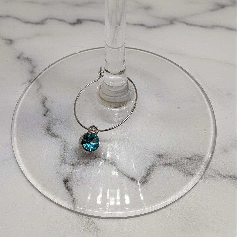 A silver-tone wine glass charm with a small, round, sparkling teal blue gemstone, shown resting on the stem of a clear glass wine glass against a white textured background.
