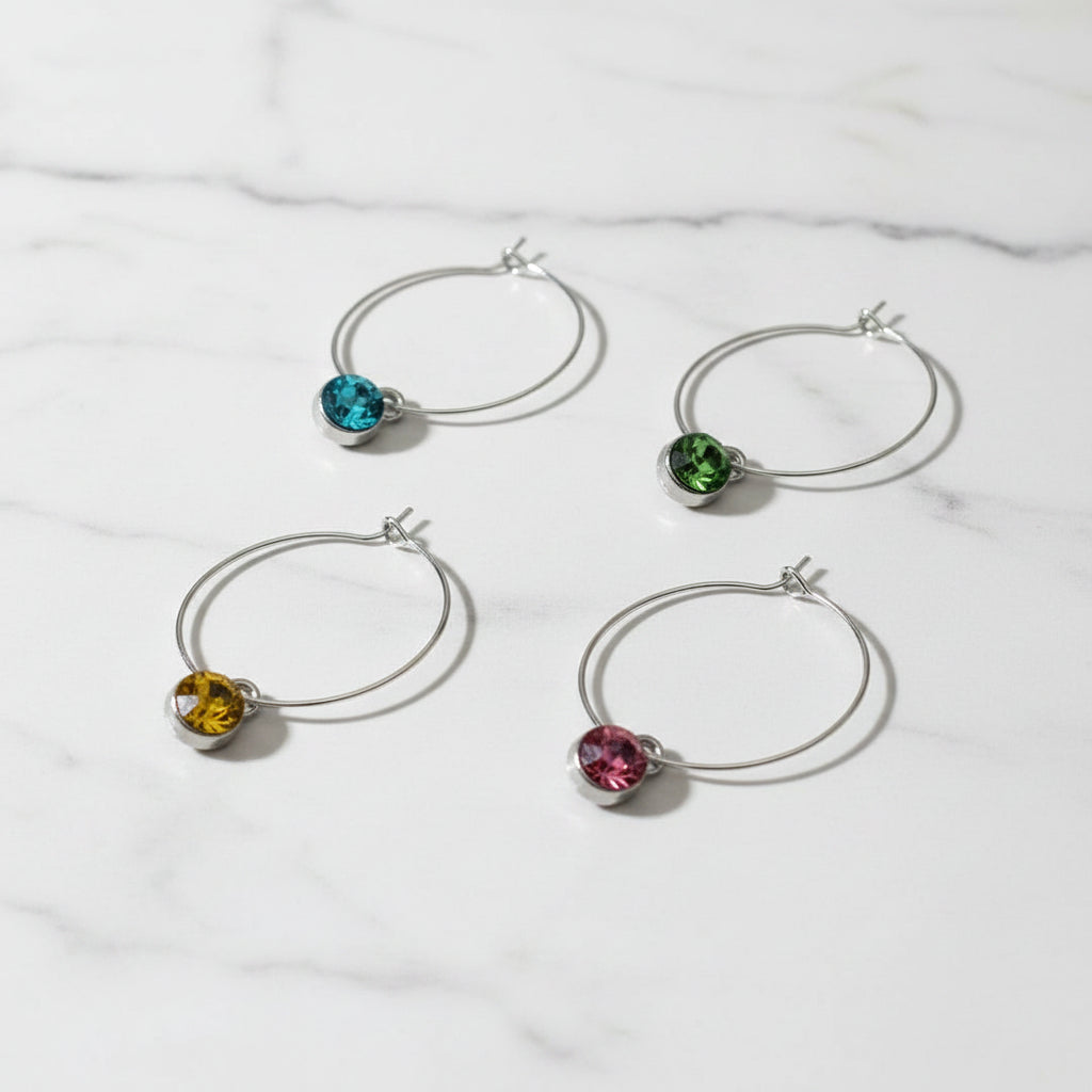Four silver hoop wine markers with round bezel-set gemstones in blue, green, yellow, and pink, arranged on a light gray textured background.