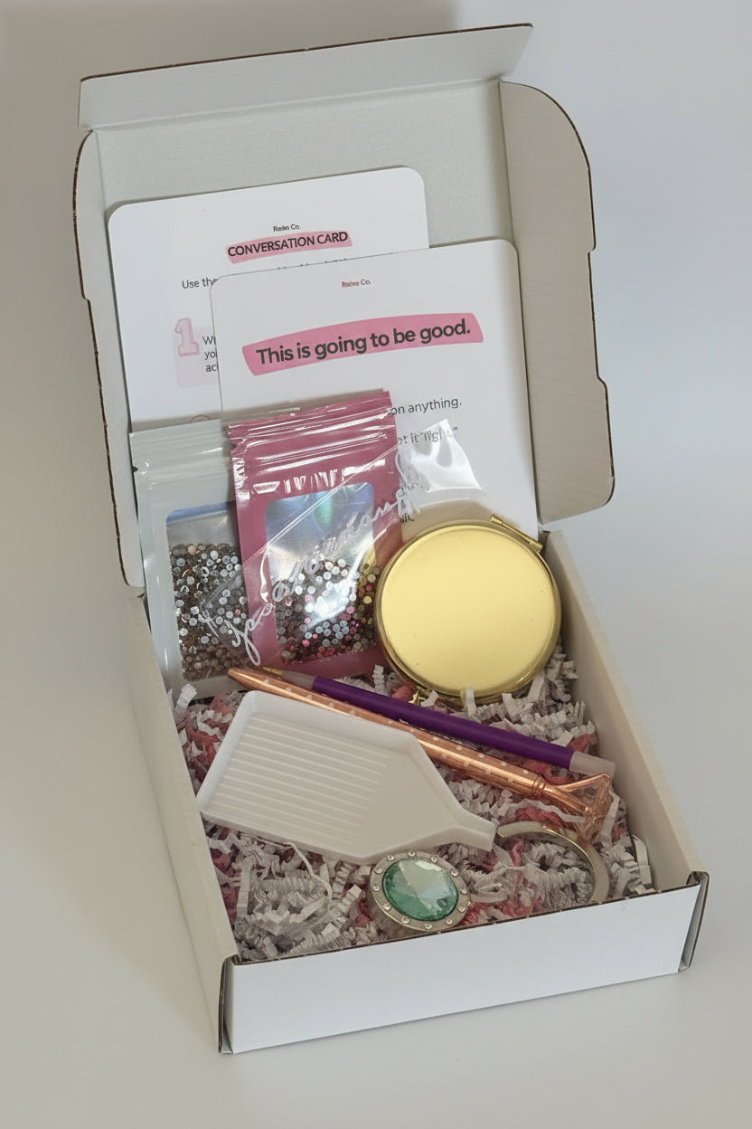 An open white gift box filled with assorted stationery-style gift items, including a gold compact, pink and champagne rhinestones in bags, a purple rhinestone picker, a green gem purse hanger, and printed cards inside shredded paper filler.