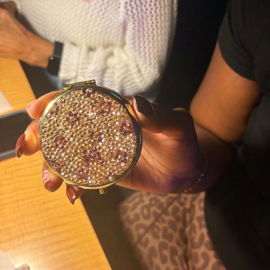 A person holds a round compact mirror covered in sparkling rhinestones with a gold-tone frame. The background shows a wooden table, a person in a white knit sweater, and leopard-print pants.