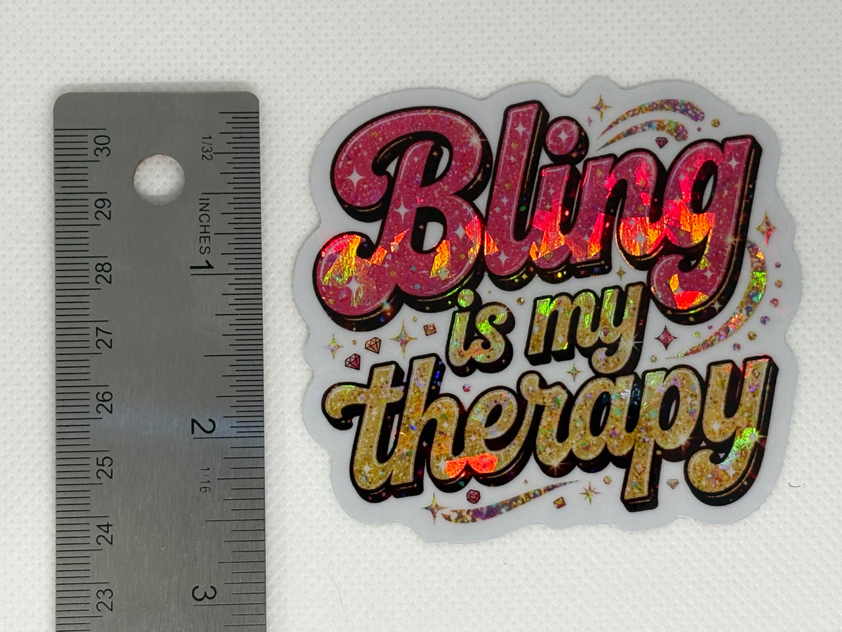 A holographic glitter sticker with the text 'Bling is my therapy' in pink, gold, and holographic lettering, shown below a metal ruler on a white textured background.