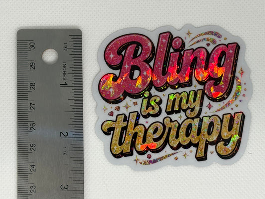 A holographic glitter sticker with the text 'Bling is my therapy' in pink, gold, and holographic lettering, shown below a metal ruler on a white textured background.