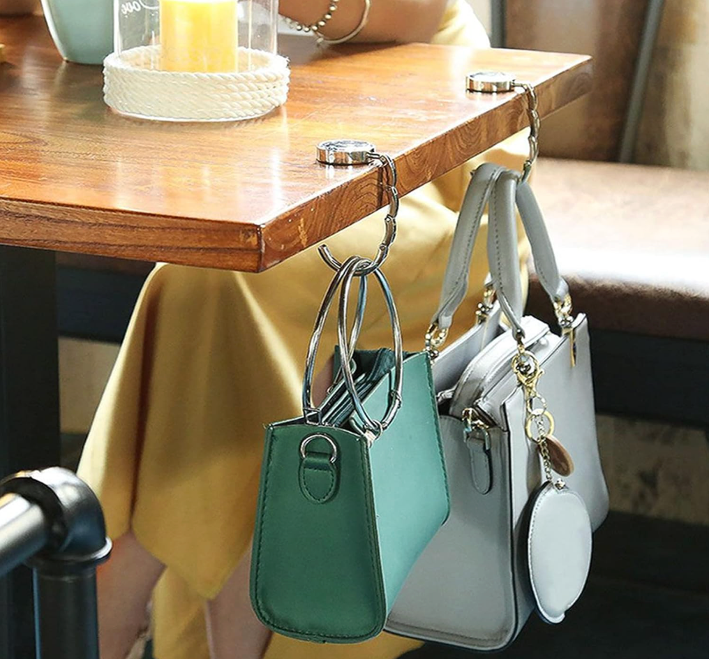 Two small structured handbags hang from metal table hooks attached to a wooden table in a real indoor setting. One bag is teal green and the other is light gray, both with silver-tone handles and hardware.