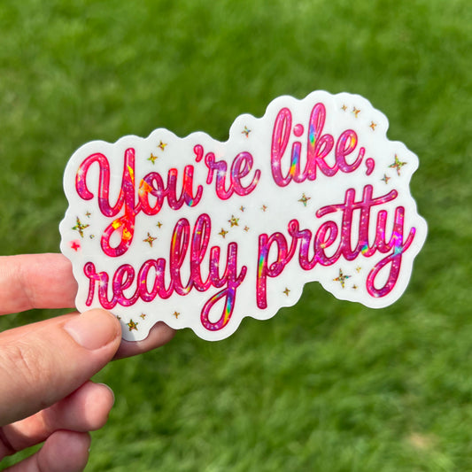 A hand holds a die-cut sticker that reads 'You're like, really pretty' in pink script with small gold stars, against a blurred green grass background.