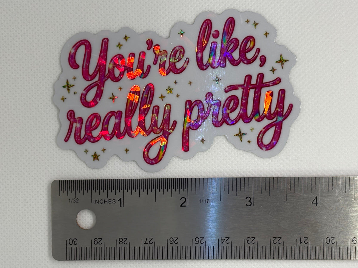 A die-cut vinyl sticker with the phrase 'You're like, really pretty' in holographic pink and purple glitter script, shown next to a metal ruler for scale on a white textured background.